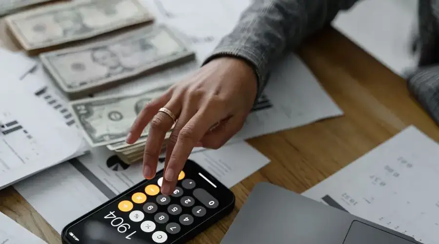 Close-up of a person using a smartphone calculator amid money and financial documents on a wooden table.