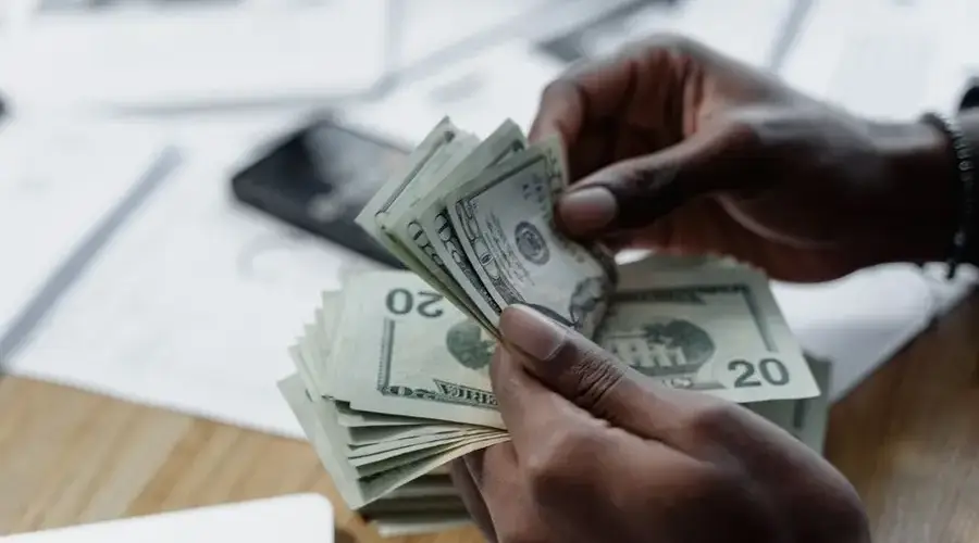 A close-up of hands counting US dollar bills on a wooden table with documents and a smartphone nearby.