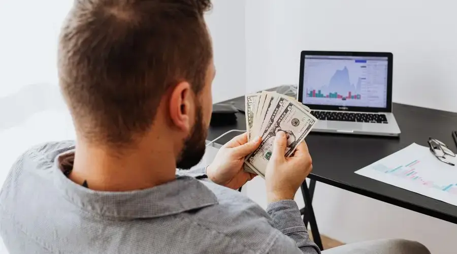 Man counting money at a desk with a laptop showing financial data.