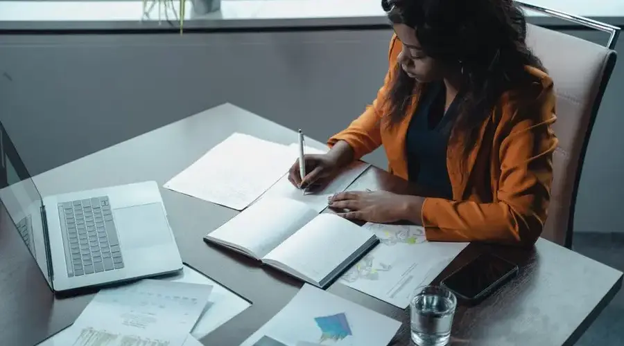 Businesswoman in office writing notes with documents and laptop, focused on analysis and productivity.