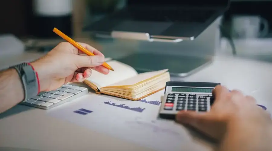 Hands using calculator and taking notes on desk with laptop, showcasing business analytics.