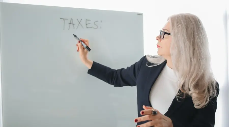 Senior woman writing on whiteboard during a tax discussion in office setting.