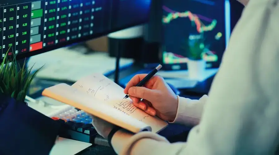 Person analyzing financial data on screens, making notes. Ideal for business and finance themes.
