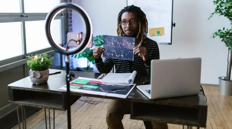 A financial vlogger in a home studio discusses cryptocurrency trends using charts and a laptop.