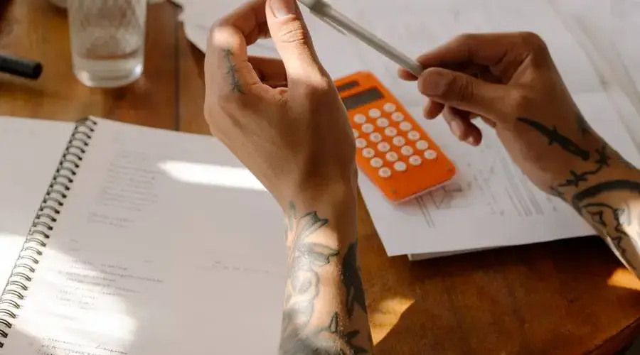 Close-up of tattooed hands holding a pen next to a calculator and notebooks.