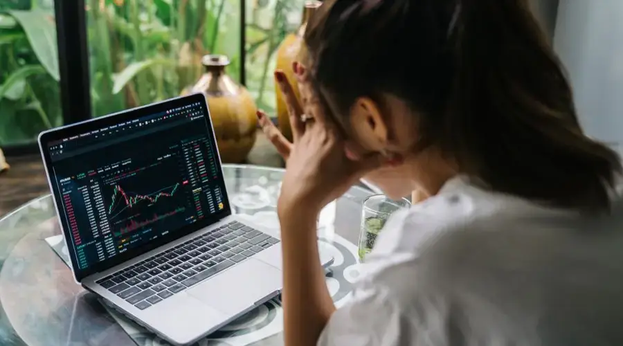 Woman analyzing cryptocurrency market charts on a laptop, looking stressed in a cozy indoor setting.
