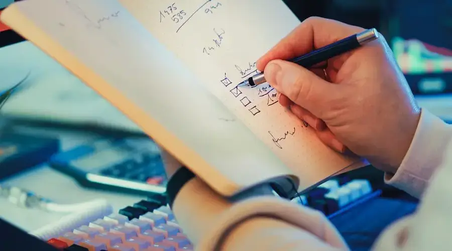 A hand writing in a notebook during stock market analysis on a desk.