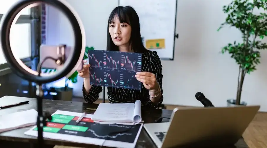 A woman vlogger discusses stock market trends using charts in a home studio.
