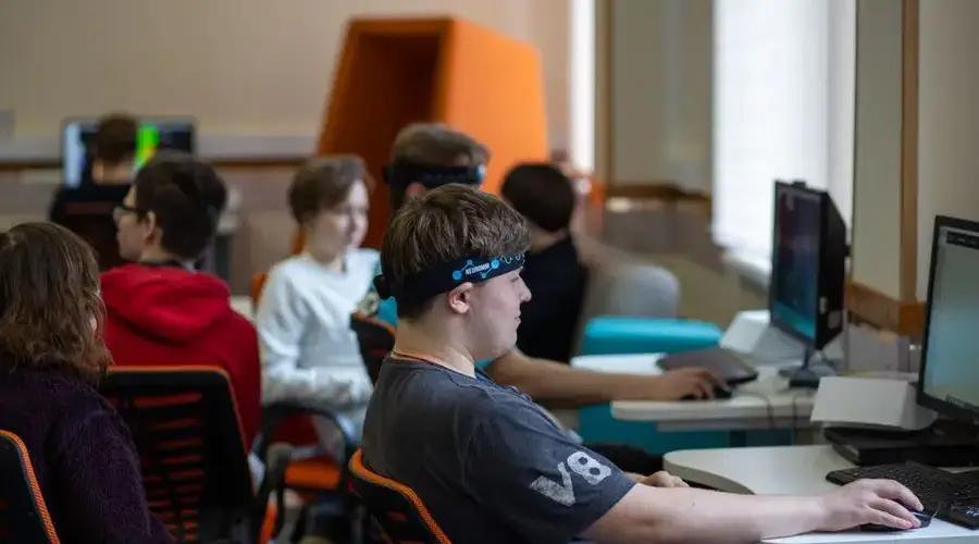 Group of teenagers engaged with computers and headsets in a modern classroom.