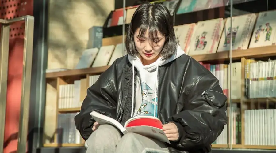 Asian woman reading outside a library, wearing a black jacket, in sunlight.