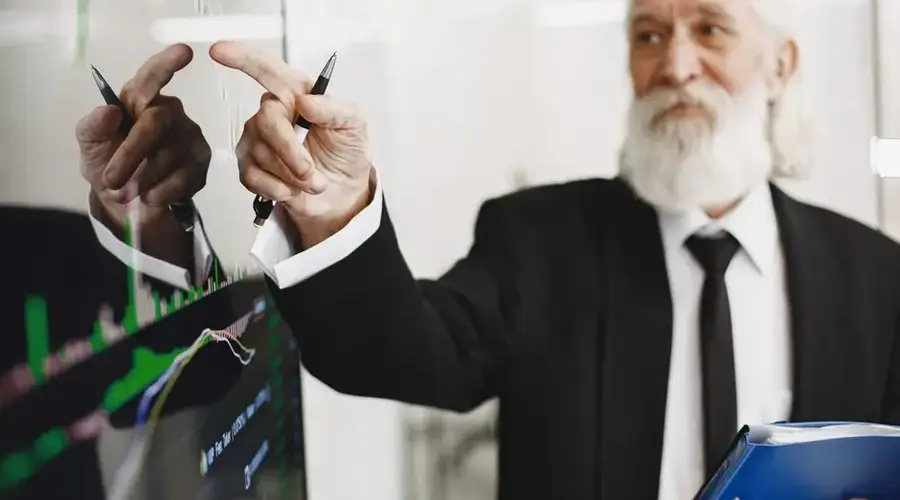 Elderly businessman with beard presenting financial trends on a screen.
