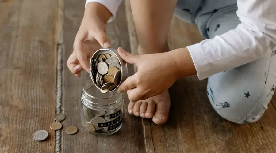 A child adds coins into a glass jar labeled for savings on a wooden floor.