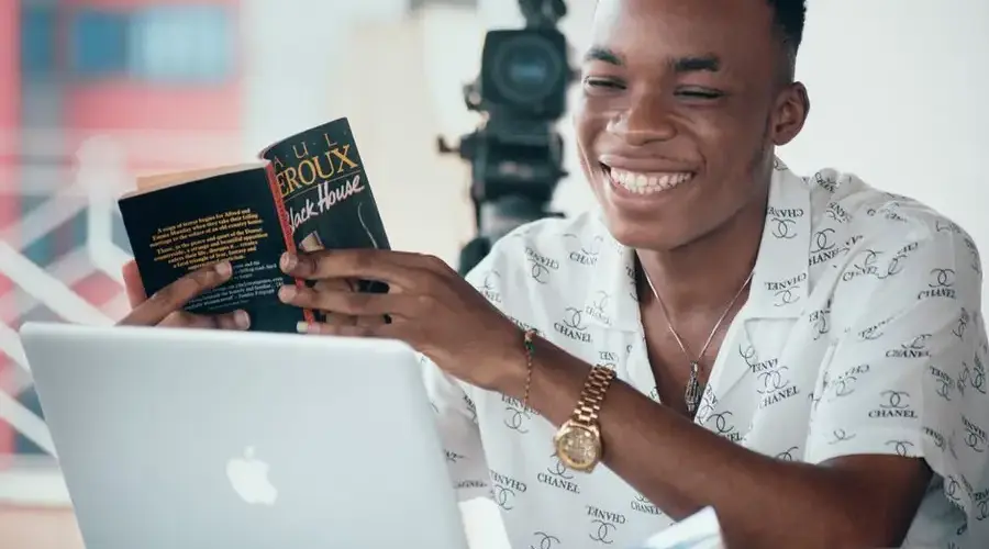 A smiling young man enjoys reading a book while using a laptop indoors, Douala, Cameroun.
