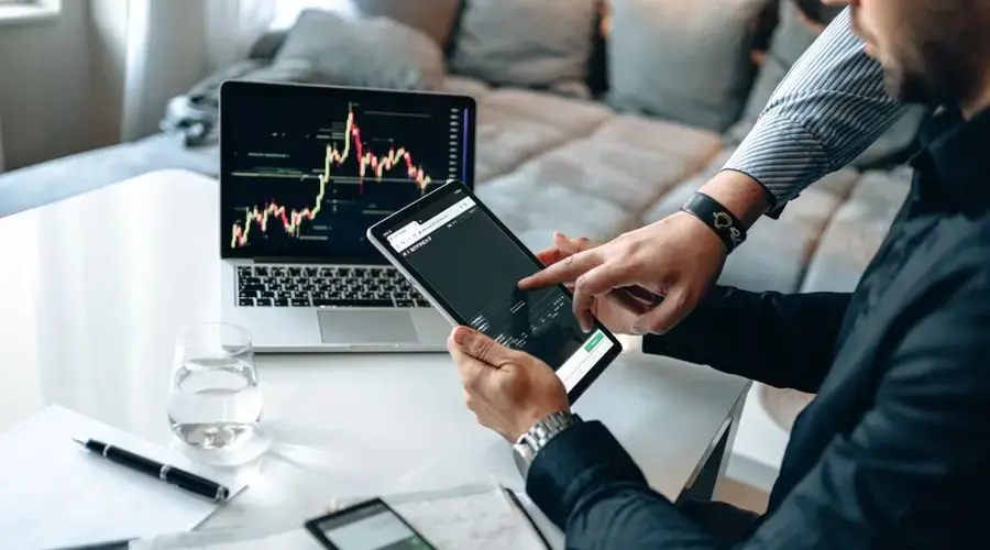 Professionals reviewing stock market trends on a tablet and laptop, discussing investment strategies.