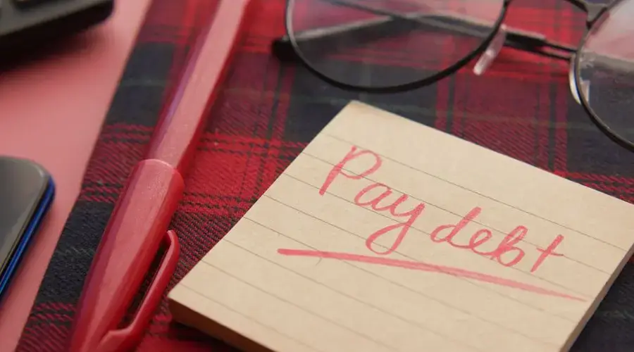 Close-up of a note reading 'Pay debt' next to a red pen on a plaid fabric, emphasizing financial reminders.