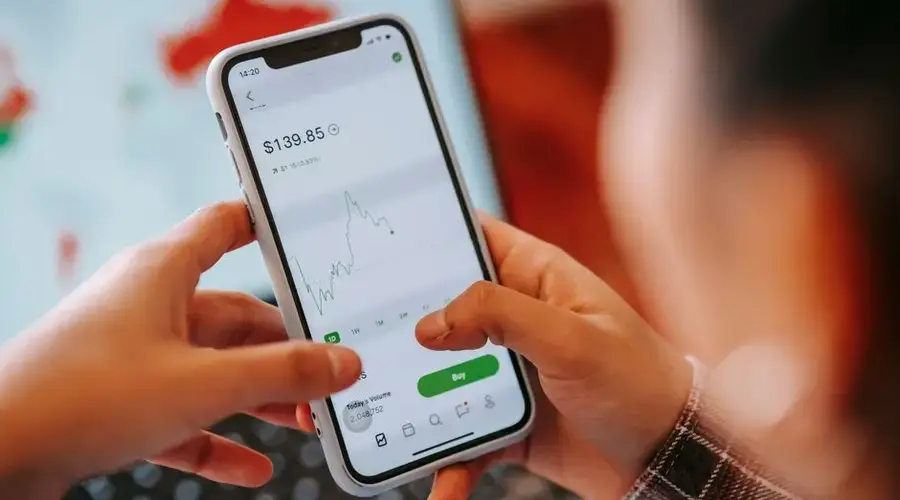 Crop anonymous trader using app on cellphone with graph and dollar price on screen in house