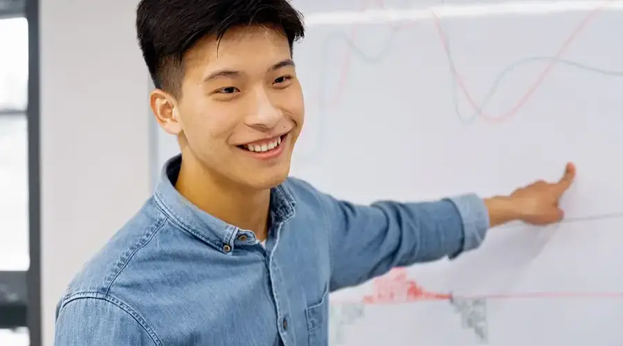 Smiling young Asian man presenting data on a whiteboard in a modern office setting.