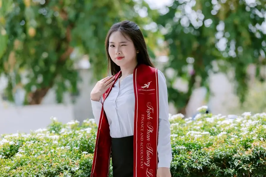 Portrait of a young graduate winking, celebrating outside, wearing a red sash.