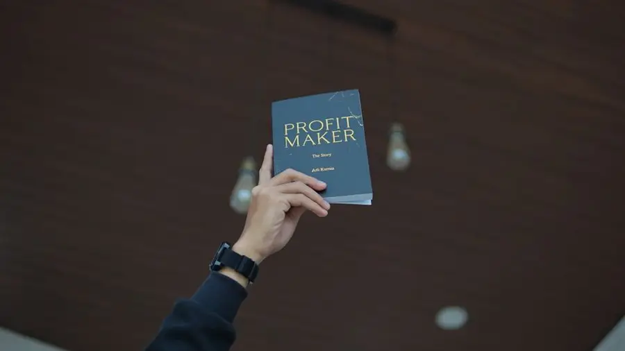 A hand raises the book 'Profit Maker' against a wooden ceiling, emphasizing motivation and success.