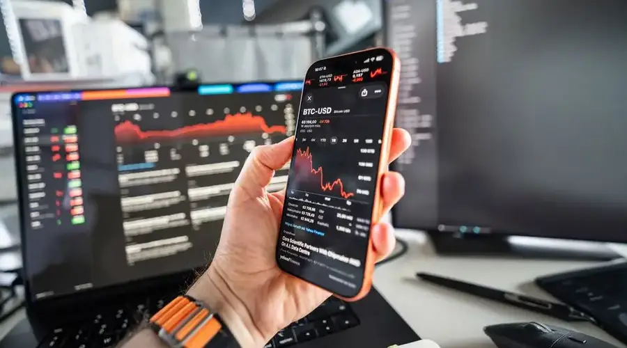 A person analyzing cryptocurrency trends on a smartphone with a declining chart on screen, set against a tech office background.