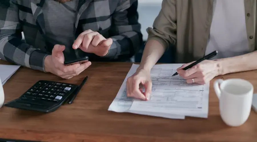 Two people working together on tax forms using a calculator at a wooden desk.