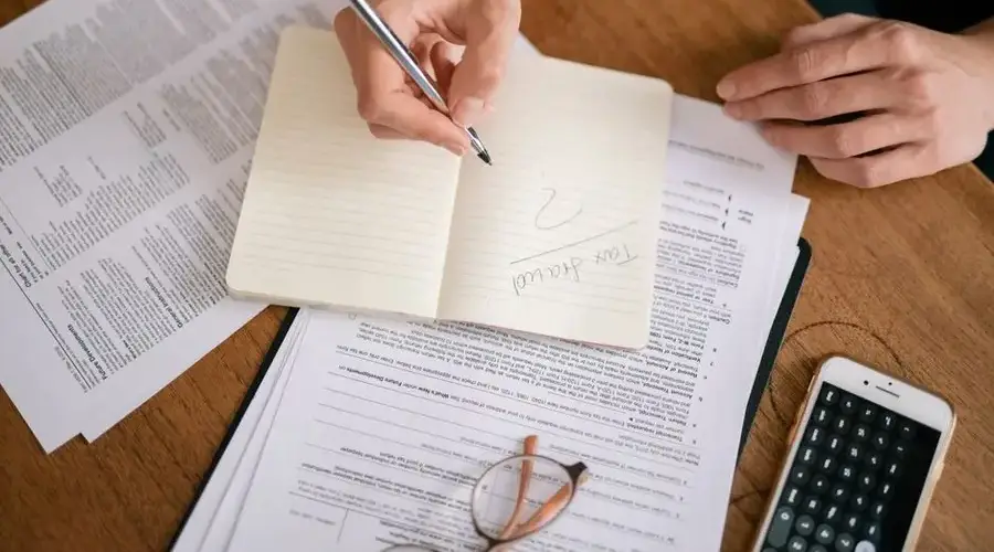 Close-up of hands writing on a notebook surrounded by tax documents and a smartphone.