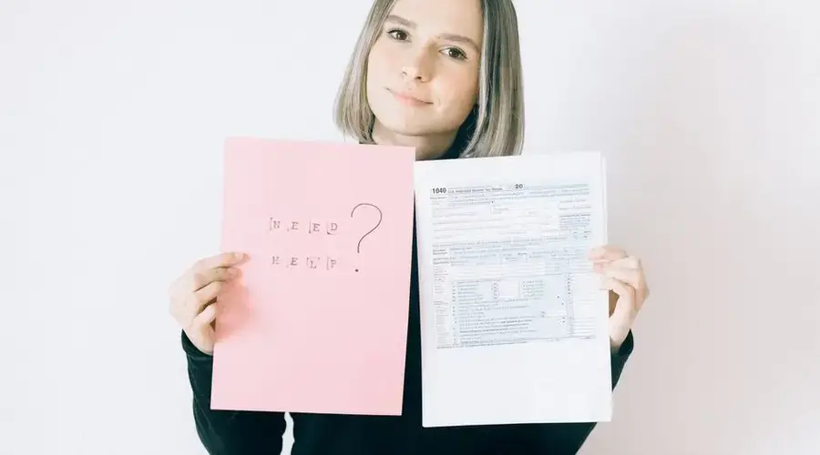 A woman in a black shirt holds tax forms and a 'Need Help?' sign, indoors.