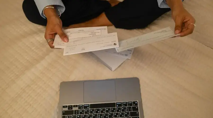 Close-up of hands holding cheques beside a laptop indoors for financial tasks.