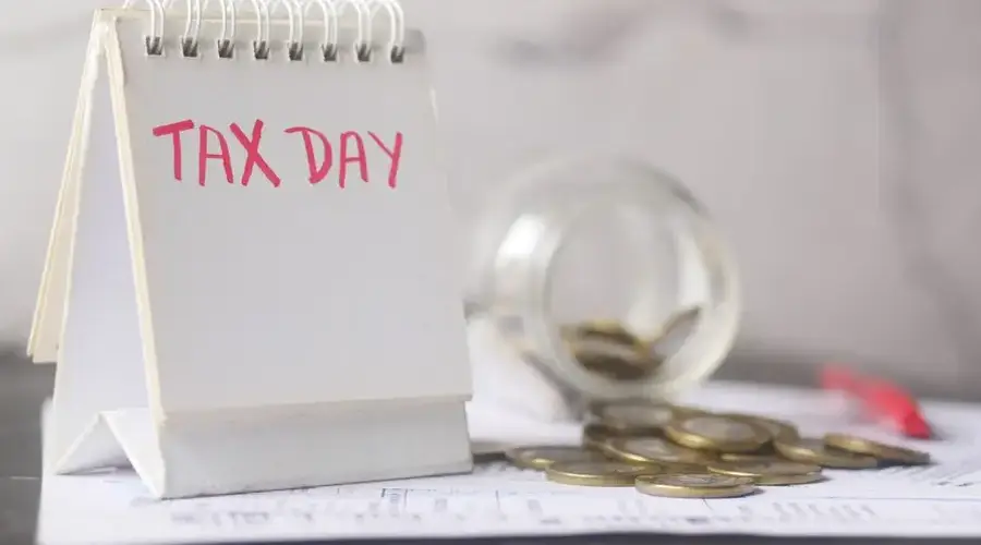 A close-up of a table calendar marked 'Tax Day' with a jar of coins and notes, symbolizing financial planning.