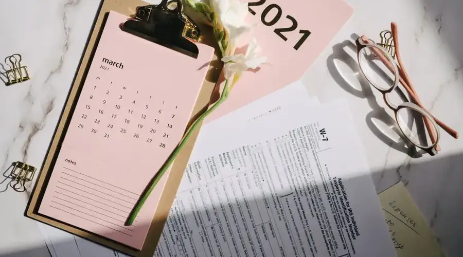 Stylish office setup with March 2021 calendar, flower, tax form, and reading glasses on desk.