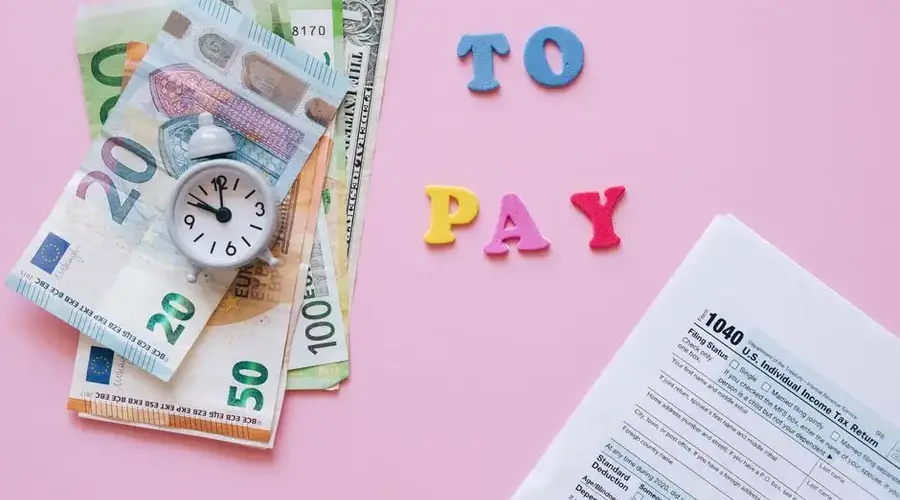 Flat lay of taxes, currency, and reminder to pay on pink background.