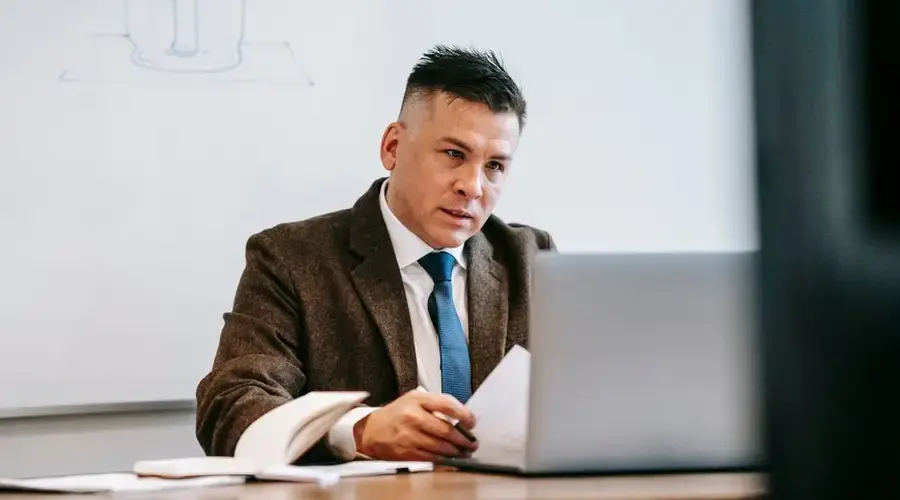 A focused professional man in a corporate setting working on a laptop with paperwork.