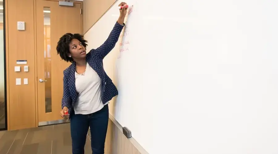 Female teacher writing on a whiteboard in a classroom, explaining a lesson to students.