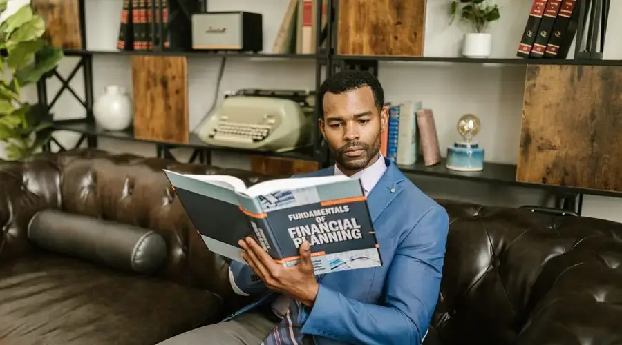 A man in a blue blazer sits on a leather couch reading a financial planning book.