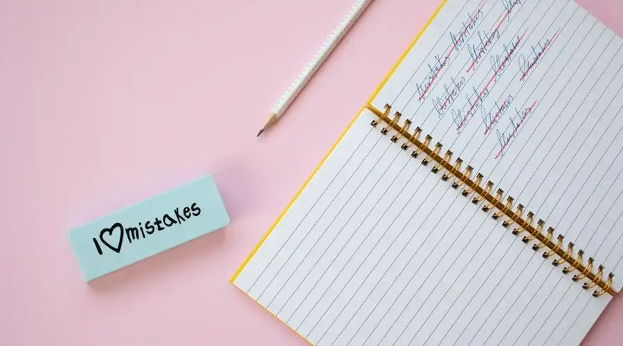 Flat lay of a spiral notebook and eraser on a pastel pink background with crossed out words.