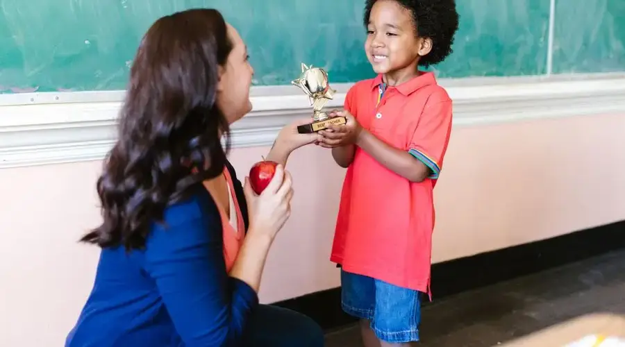 A teacher presents a trophy to a smiling child in a classroom setting.