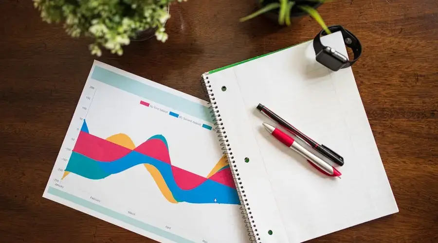 Top view of a workspace with graph, notebook, pens, and smart watch.