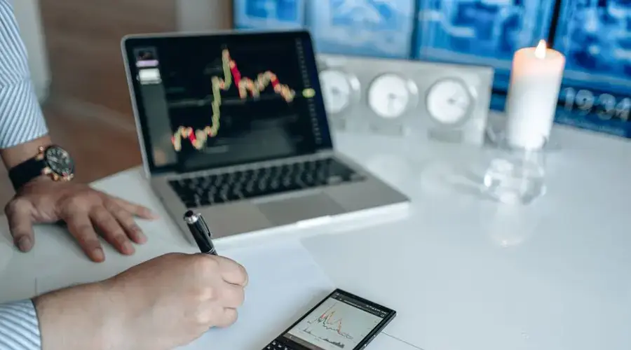 Business analyst writing with laptop and smartphone showing trend graphs on a white desk.