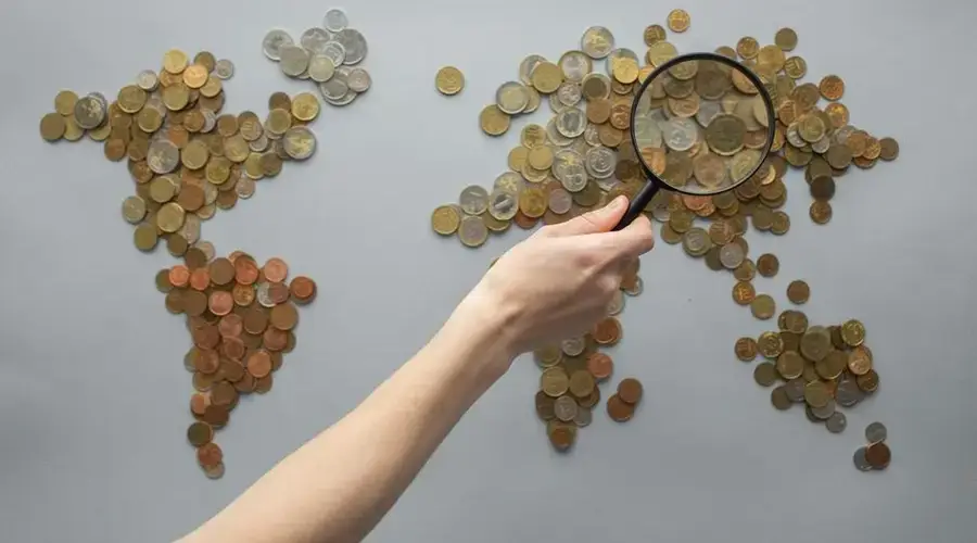 Top view of crop unrecognizable traveler with magnifying glass standing over world map made of various coins on gray background