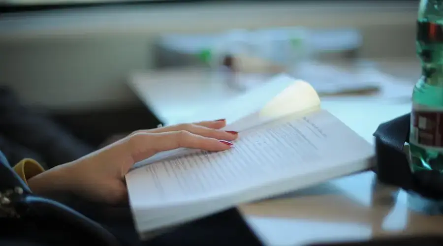 A woman with manicured nails is reading a book at a desk, emphasizing study and relaxation.
