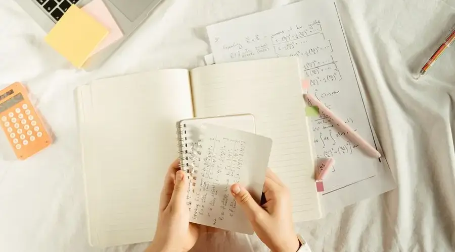 Hands holding a notebook with math equations, calculator, and laptop on a study desk.