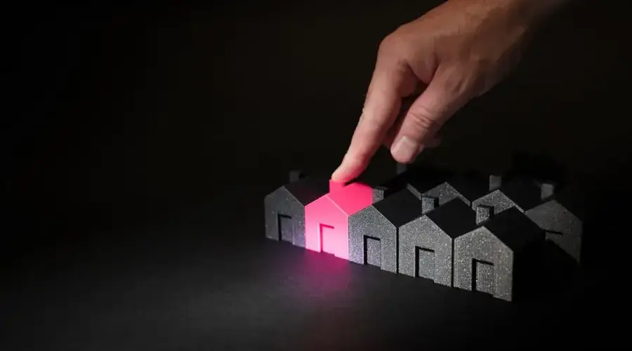 A hand selecting a glowing pink house among dark miniature houses, symbolizing real estate choices.