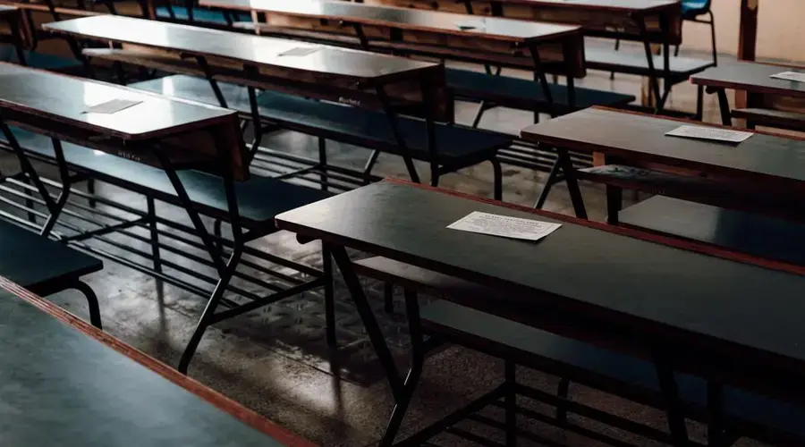 A row of unoccupied desks and benches in a classroom with exam papers on top.