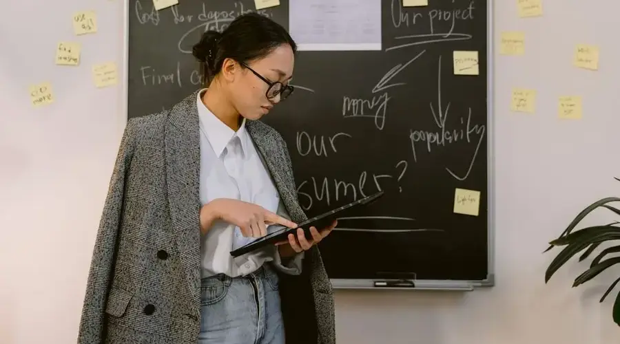 Asian woman in a gray blazer holds a tablet near a blackboard with notes and sticky notes in an office setting.