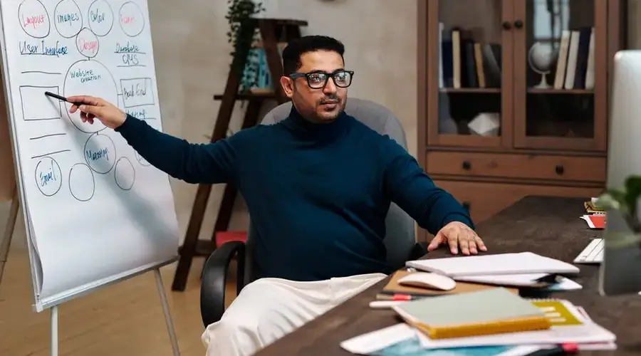 Man teaching online, pointing at a flipchart in a cozy office space. Perfect for online education themes.