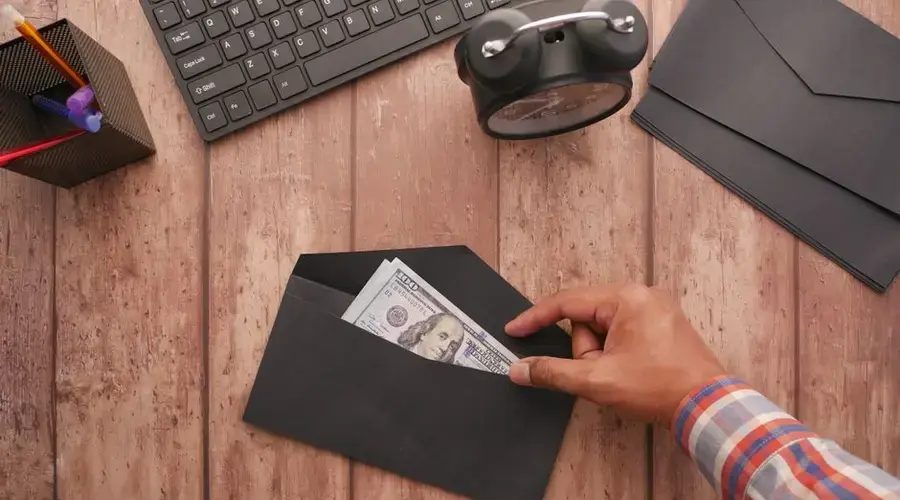 A hand placing a 100 dollar bill in a black envelope on a wooden desk with stationery, keyboard, and alarm clock.