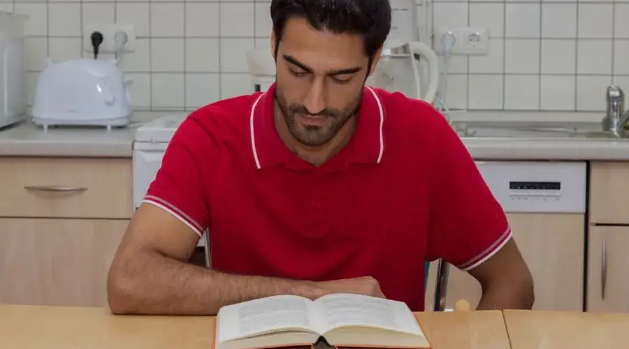 A man in a red polo shirt reading a book at a kitchen table, focused and engaged.