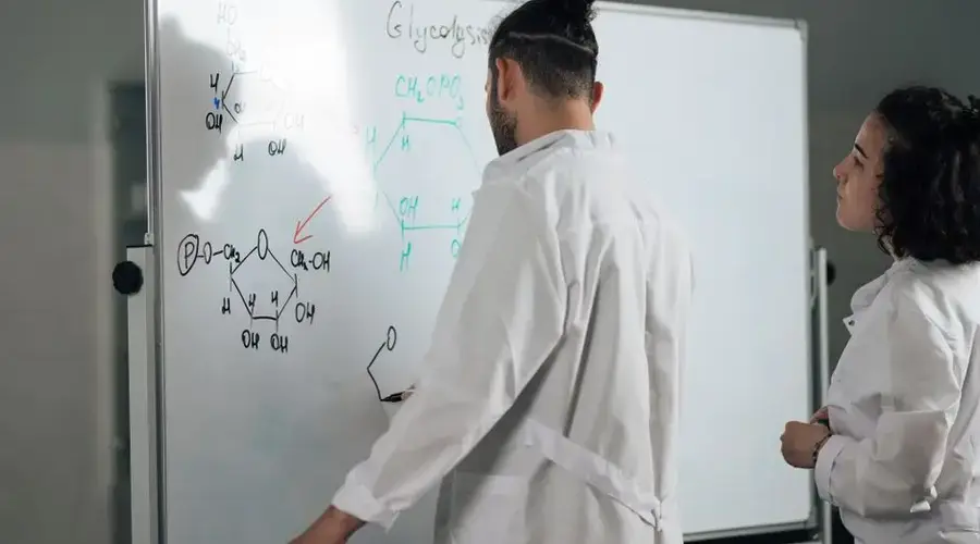 Two scientists in lab coats writing and discussing chemical formulas on a whiteboard in a laboratory setting.