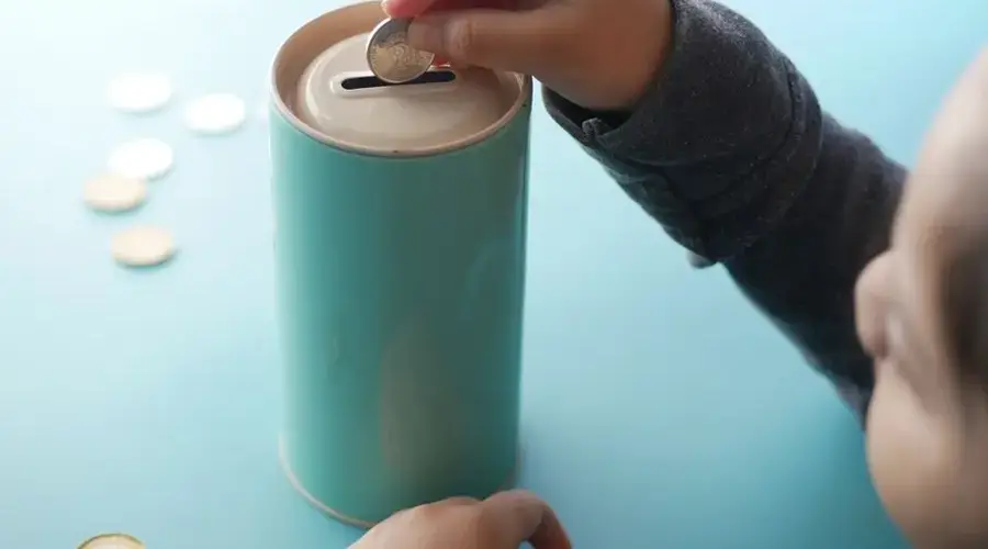 A child placing a coin into a teal piggy bank, representing saving money and financial education.