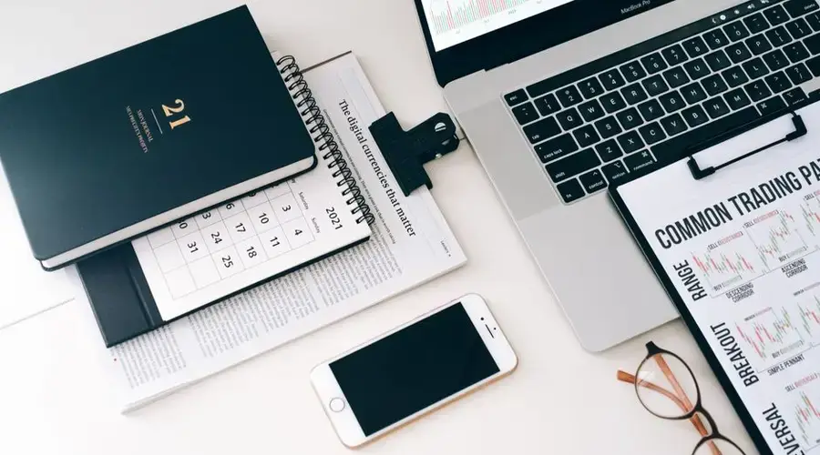 Flat lay of a modern workspace with a laptop, smartphone, planner, and trading charts on a white surface.